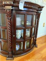 View of front curved glass door with wood framing and carved wooden columns on the sides, showing china dishes inside.