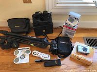 Photo showing collection of electronics including Canon camera, two pairs of binoculars (Bushnell and Jason Empire), Velbon tripod, Omron blood pressure monitor, Omron pedometer with manuals, USB cable, and carrying bag on wooden floor.