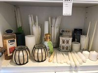 Shelf with assortment of white taper and pillar candles, two candles in glass holders with black metal frames, small gold oil lantern, and fragrance oil bottle.