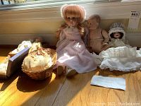 The seven antique and vintage bisque dolls displayed on a wooden floor near a window. One large doll wears a pink dress and bonnet; others are smaller with various clothing and accessories including one in a basket and one with a bonnet.