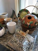 Several flower pots white, brown terracotta, purple small pots with African violets, small decorative sheep garden figure, a green metal triangular birdhouse, painted gourd birdhouse and a bag of birdseed on countertop.