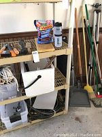 Photo of shelving unit with various garden tools including shovels, rakes, broom, coiled garden hose, bag of fertilizer, white bucket with rope inside, and floor mat on bottom shelf.
