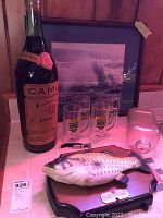 Display of a large Camus Armagnac bottle, two Collins Pub mugs, framed picture of ocean waves, clear glass jar, Big Mouth Billy Bass fish decoration on wood plaque, and a corkscrew bottle opener on white cloth surface against wood panel background.