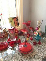 Photo showing a variety of hummingbird feeders and parts arranged on a granite counter near a window. Visible are several red and clear glass feeders, a white ceramic flowered piece, and other decorative items.