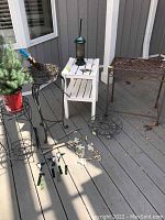 Overview of lot showing iron patio table, two metal plant stands (one with a small evergreen), small white wooden side table with bird feeder, glass wind chimes broken on ground, and plant wheels.