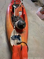 Full view of the orange Swifty kayak showing paddle, buoy, gloves, and sealant container placed on top inside a basement setting.