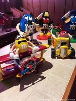 Overview of the lot showing multiple M&M's figurines and candy dispensers arranged on a table in a basement bar setting. Notable pieces include yellow M&M on an airplane, blue M&M with saxophone, red fireman M&M, and red and green M&Ms in a yellow hot rod car.