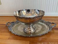 Wide view of the silver-tone punch bowl centered on a matching ornate oval tray, placed on wooden floor
