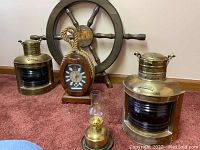 Full view of set including ship wheel, two brass oil lanterns, the rope-handled quartz clock, and a small brass and glass oil lamp on carpeted floor.
