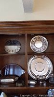 Cabinet display of silver plate pieces including round trays with cut-out designs, oval serving dishes, small coasters, and footed bowl