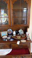 Photo showing 16 decorative items including plates, a carved bird, vessels, a gnome figurine, a framed painting, and more arranged on a wooden sideboard.