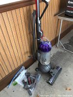 Full view of Dyson DC65 Animal vacuum standing against the wall with power cord, clear dust bin, ball base, and attachments in plastic bag beside it on floor.