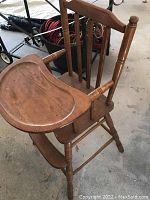 Photo showing wooden vintage highchair with curved tray attached, angled from side. Shows turned spindle legs and backrest with vertical slats.