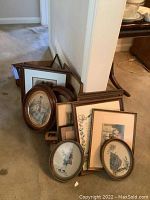 A collection of nine framed art prints and needleworks stacked on floor with wooden frames, including oval and rectangular frames.