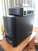 Black Bose Acoustimass Module subwoofer on a wooden table with other items partially visible.