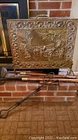 Full view of ornate brass-like fireplace screen set on brick hearth showing embossed pastoral scene and fireplace tools propped in front.