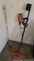 Two fishing rods with attached reel and a Treasuremaster metal detector with headphones and orange base coil, standing against a wall.