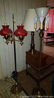 Photo showing three lamps: one brass table lamp with fabric shade and two brass floor lamps including a double light with red glass shades and a four-light lamp with exposed bulbs