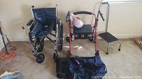 Shows the four main items in the lot; folding wheelchair, red walker, black stepstool and navy wheeled duffel bag on carpeted floor.
