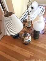 Overview of five lamps, three ceramic and two metal, arranged on wooden floor; includes pleated shade and hobnail milk glass lamp