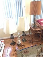 Four lamps grouped on a beige carpet in front of window with curtains, showing two ceramic lamps with square white shades and one brass lamp with pleated shade, one lamp base without shade.
