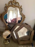 Large ornate gold carved wall mirror with intricate floral and scroll patterns. Two rustic wood framed hexagonal mirrors and a brass-toned small standing vanity mirror are in front.