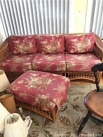 Front view of rattan sofa with three red floral seat and back cushions and matching ottoman in front