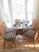 Photo showing the square rattan table with glass top and four rattan chairs with plaid padded seats arranged around it near a window.