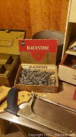 Box of Blackstone nails along with a hand saw shown on a wooden surface, with other items in the background