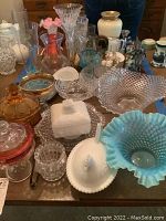 Overview of table with assorted glassware and decorative items including blue hobnail bowl, milk glass dishes, etched vases, crystal bowl, amber candy dish, and ceramic pitcher.