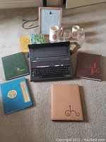 Group photo showing four yearbooks of varied colors including blue 'Challenger' embossed cover, black Brother AX-24 typewriter, and four pieces of college glassware with logos. Items arranged on carpeted floor.