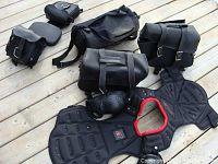 Overview of two sets of motorcycle saddle bags in black leather and nylon, and body armour vest laid out on wooden surface.