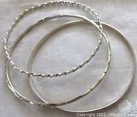 Three different vintage sterling silver bangles photographed together on white fabric background showing their shapes and textures.