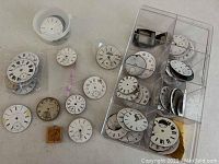 Photo showing multiple antique pocket watch faces and parts laid out in plastic containers and bags. Faces have Roman and Arabic numerals, some with wear and tear.