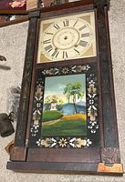 Full view of antique wooden wall clock showing upper clock face with Roman numerals and lower painted glass panel with landscape scene and decorative floral designs on sides.
