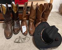 Photo showing three pairs of brown cowboy boots, a black cowboy hat, and a pair of metal stirrups arranged on carpet.