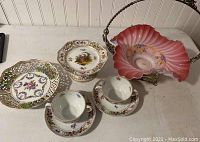 Full lot view showing Dresden pedestal plate, two creamed soup cups and saucers, and Fenton style pink brides basket with silverplate handle and base