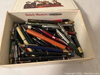 Box full of diverse vintage fountain pens and pencils, mixed colors and styles visible.
