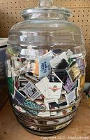 Front view of large clear glass jar filled with vintage matchbooks showing multiple matchbook logos inside, viewed against pegboard background.