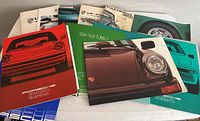 Photo showing a collection of Porsche and Volkswagen brochures spread out, featuring colorful covers with Porsche model images like 911 SC Turbo, 924 Turbo, and VW 2000.