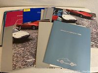 Photo showing multiple Porsche factory car brochures laid out overlapping on a flat surface, covers include various Porsche sports cars with the Porsche logo.