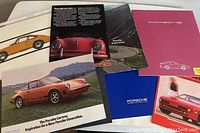 Photo showing multiple Porsche brochures with covers visible including a red car, a yellow car illustration, and blue folder.