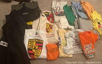 Overview of folded Porsche parade and car event shirts in multiple colors with various Porsche logos and graphics, laid out on floor.