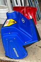 Close-up side view of electric snow shovel unit showing blue motor housing, Snow Joe logo, and red snow shovel blade with black edge.