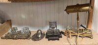 Overview of four desk accessories on a shelf: ornate brass double inkwell, silver horseshoe inkwell, black cast iron double inkwell, brass desk lamp