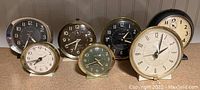 Seven vintage wind up alarm clocks including Westclox Big Ben and Baby Ben models, assorted colors and sizes shown on shelf.