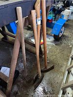 Four heavy tools: three wood handle axes and one wood handle sledge hammer shown standing on concrete floor against metal workbench.