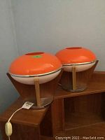Two orange plastic shade bedside lamps with teak veneer bases and metal circular stands shown on wooden furniture.
