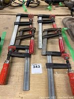 Six adjustable bar clamps with red wooden handles laid on a wooden surface.