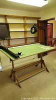 Drafting machine on wooden trestle table in room with shelving. Shows green drawing surface with mechanical arms and magnifying lamp on left side.